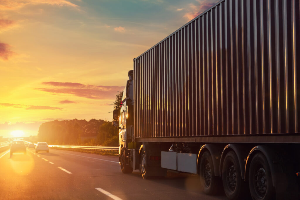 Scenic back view big long heavy semi-treailer truck with sea shipping container driving highway dramatic warm morning evening sunrise sun sky sunset. Cargo transport industry background concept.